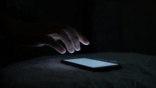 暗がりで光るスマートフォンにためらいながら手を伸ばす様子